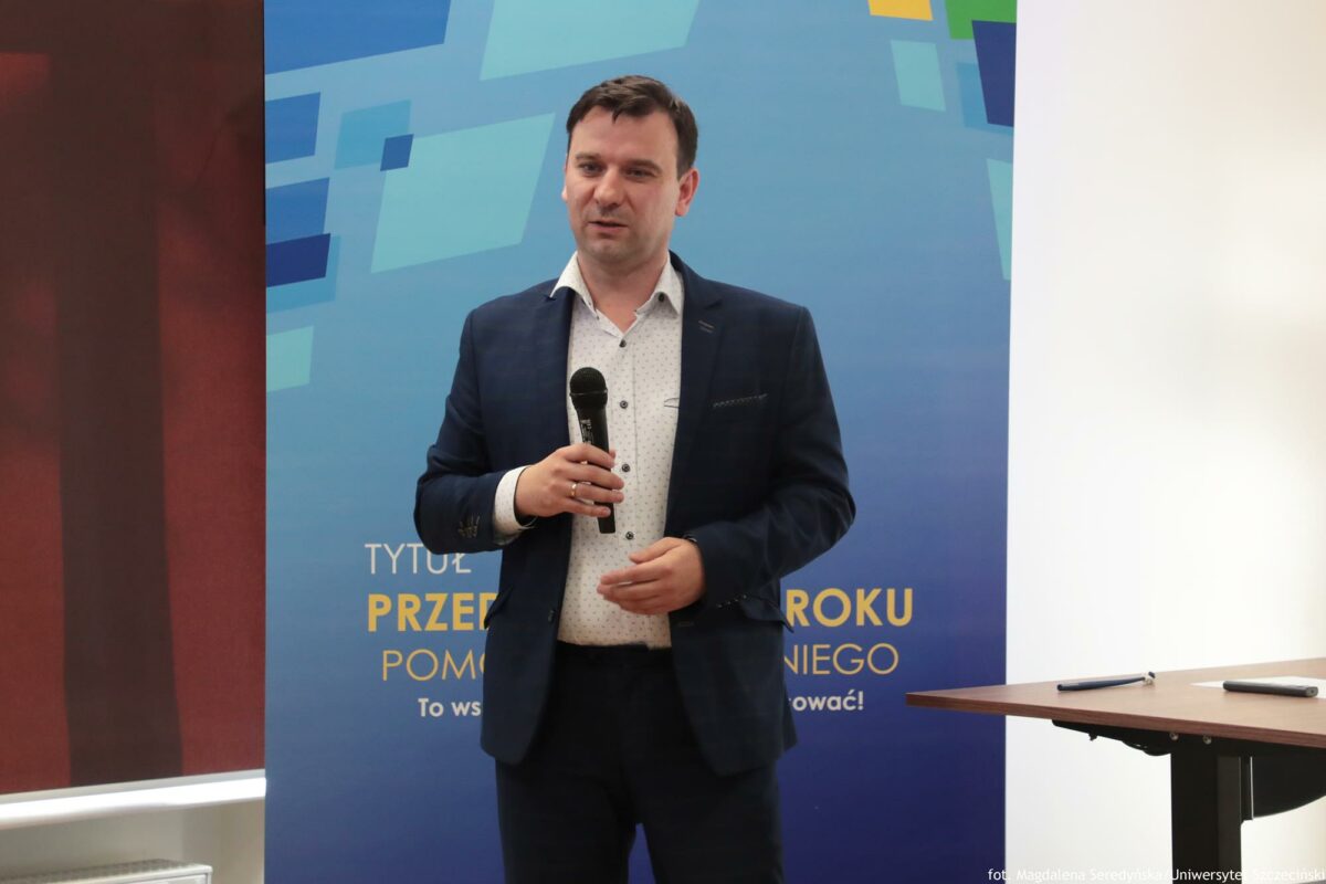 Prelegent trzymający mikrofon (ujęcie do kolan). Za nim rollup Północnej Izby Gospodarczej.