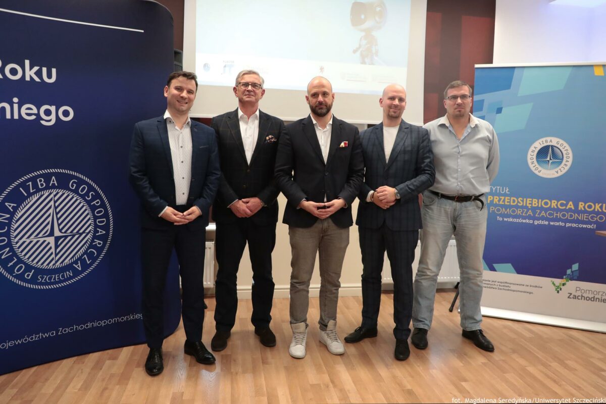 Grupa uczestników na konferencji o AI (5 mężczyzn). Za nimi na środku ekran, po prawej rollup Północnej Izby Gospodarczej, po lewej granatowa ścianka Północnej Izby Gospodarczej.