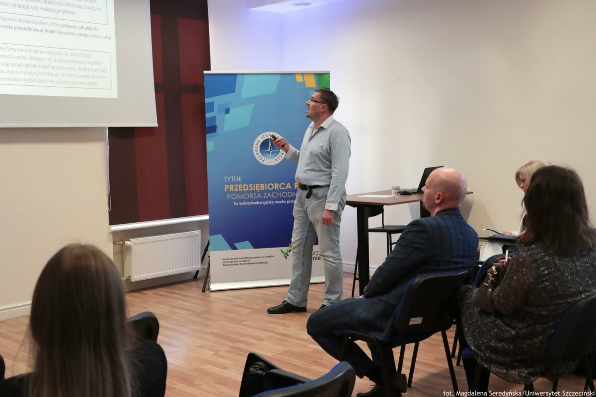 Prelegent omawia swoją prezentację, trzymając pilot i patrząc w ekran. Za nim biurko z laptopem, obok rollup Północnej Izby Gospodarczej. Na pierwszym planie słuchacze bokiem lub tyłem do kadru.