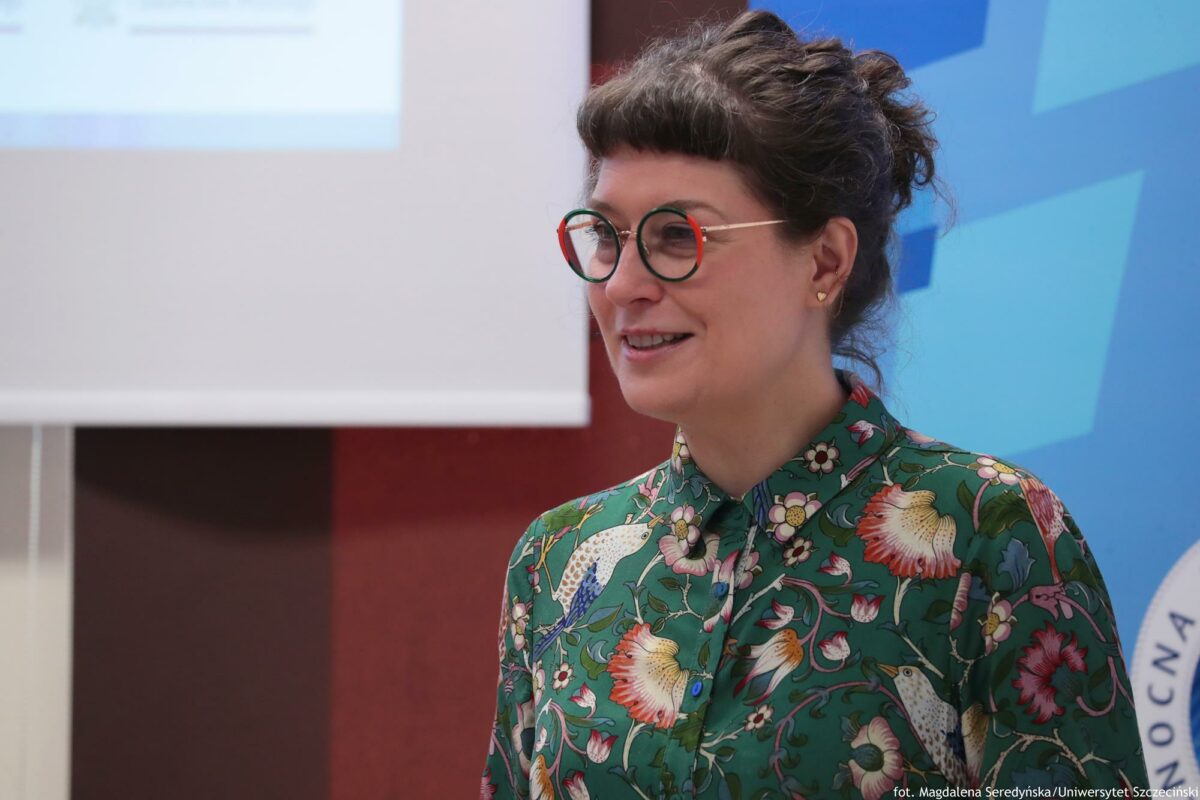 Prelegentka (ujęcie do piersi – bokiem). W tle ekran i rollup.