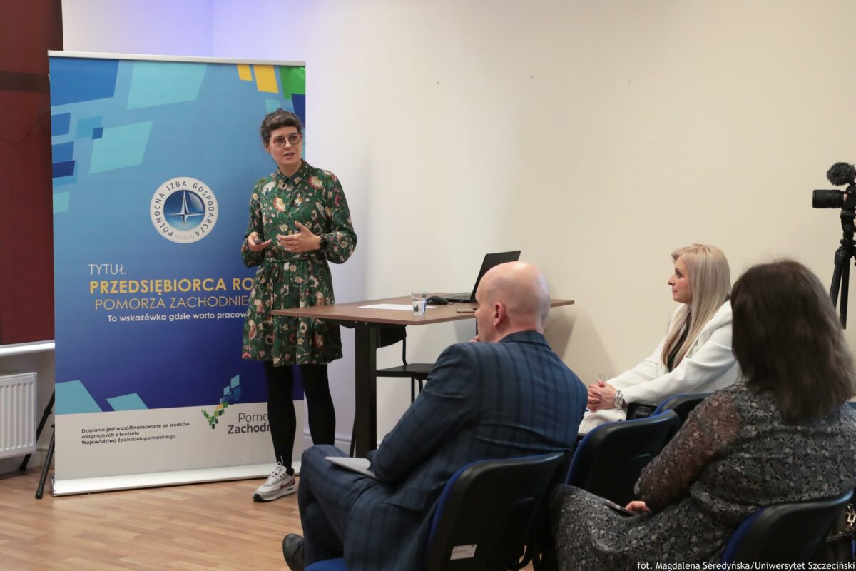 Prelegentka. Przed nią biurko z laptopem. Za nią rollup Północnej Izby Gospodarczej. Na pierwszym planie (po prawej) słuchacze tyłem do kadru.