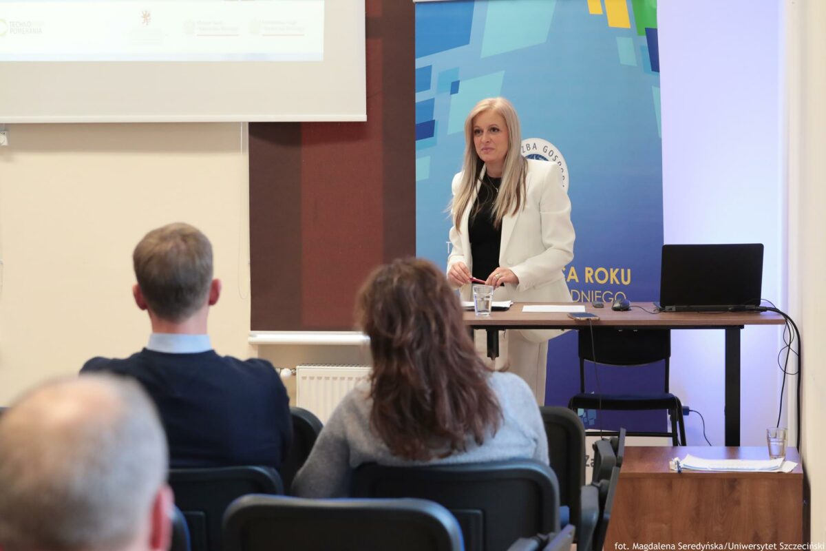 Prelegentka. Przed nią biurko z laptopem (prawa strona). Za nią rollup Północnej Izby Gospodarczej (lewa strona). Na pierwszym planie słuchacze tyłem do kadru.