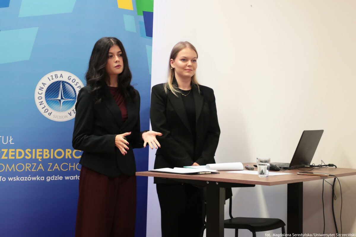 Prelegentki (ujęcie do kolan – bokiem). Przed nimi biurko z laptopem. Za nimi rollup Północnej Izby Gospodarczej.