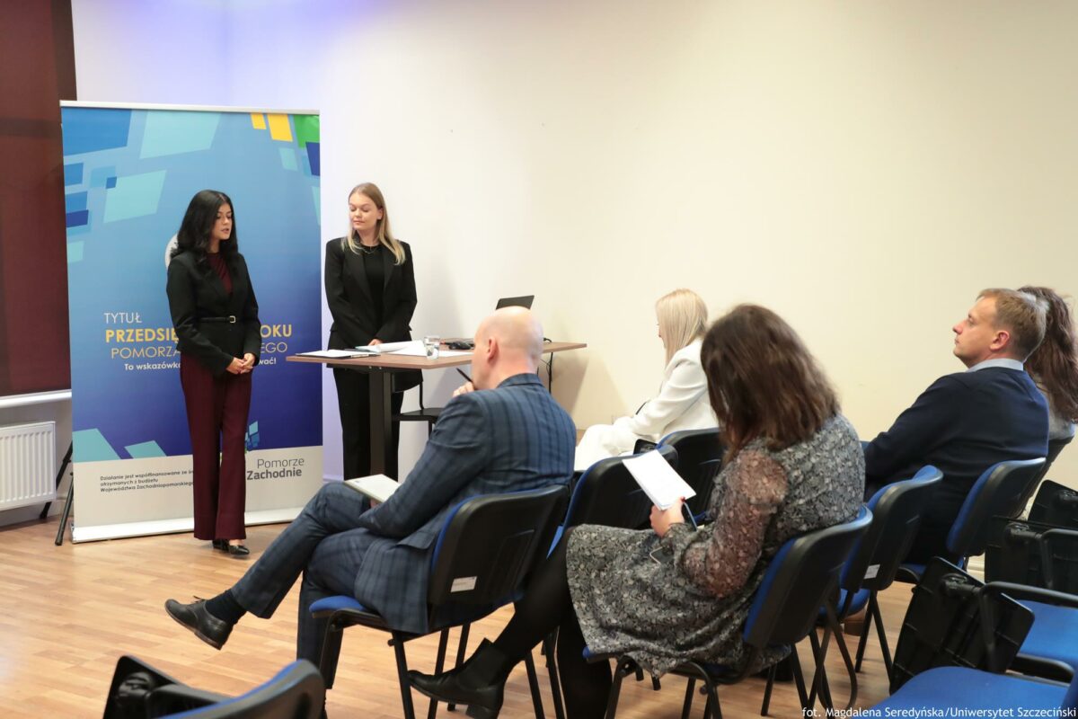 Prelegentki (ujęcie bokiem). Przed nimi biurko z laptopem. Za nimi rollup Północnej Izby Gospodarczej. Na pierwszym planie słuchacze tyłem do kadru.