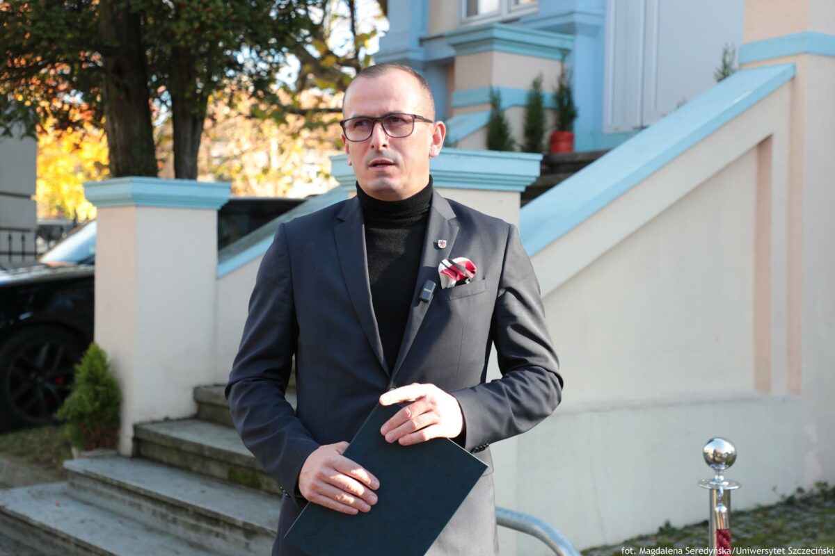 Trzymający teczkę prelegent konferencji o AI na tle schodów prowadzących do zabytkowej willi.