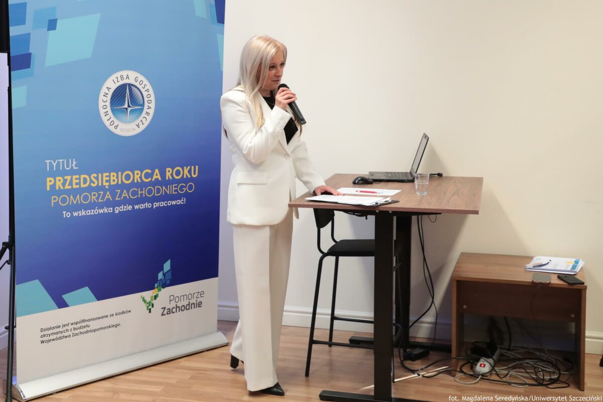 Prelegentka (ujęcie bokiem). Przed nią biurko z laptopem (prawa strona). Za nią rollup Północnej Izby Gospodarczej (lewa strona).