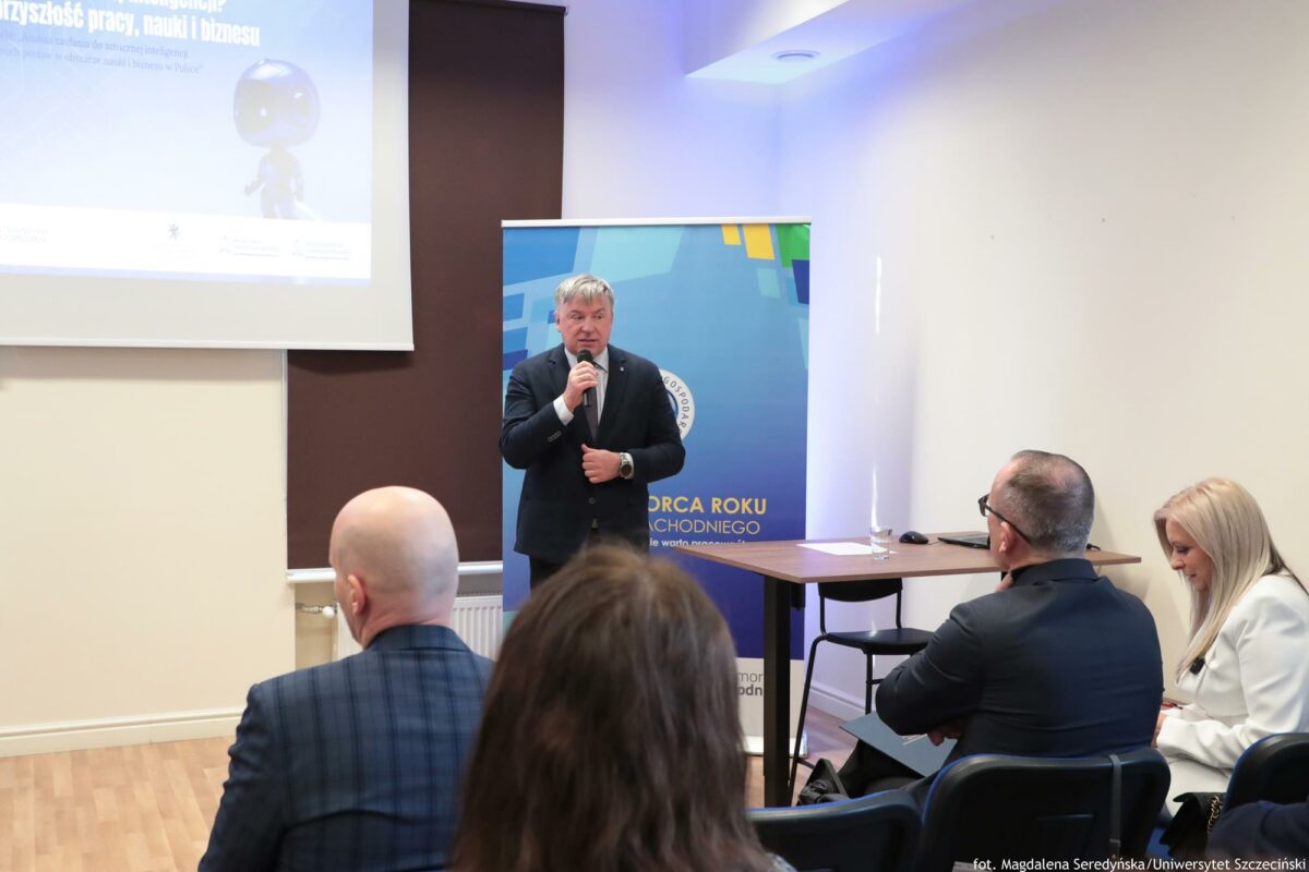 Prorektor ds. nauki Uniwersytetu Szczecińskiego otwierający konferencję o AI (ujęcie bokiem). Za nim rollup Północnej Izby Gospodarczej i ekran z prezentacją. Na pierwszym planie słuchacze tyłem do kadru.