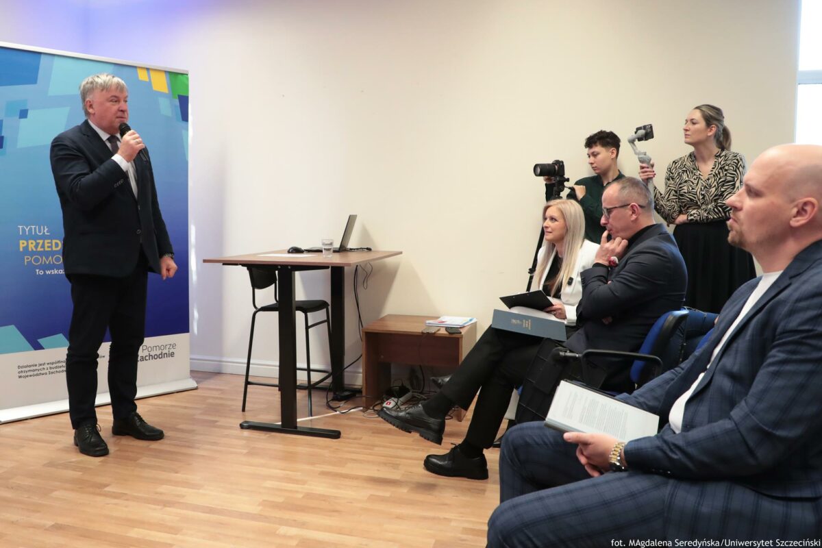 Prorektor ds. nauki Uniwersytetu Szczecińskiego otwierający konferencję o AI (ujęcie bokiem). Za nim rollup Północnej Izby Gospodarczej. Po lewej słuchacze i media.