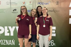 Grupa czterech młodych kobiet pozujących do zdjęcia na tle ścianki sponsorskiej na zawodach sportowych. Trzy stoją z przodu w bordowych koszulkach i z medalami na szyi; dwie z nich trzymają medale uniesione w dłoni. Z tyłu stoi czwarta kobieta, z rękami opartymi na ramionach dwóch osób z przodu. Osoba z lewej z przodu ma jasną karnację, długie jasnobrązowe włosy i czarne okulary; ma bordowe krótkie spodenki. Środkowa z przodu ma jasną karnację i falowane jasnobrązowe włosy do ramion; ma ciemne krótkie spodenki. Osoba z prawej z przodu ma jasną karnację, ciemne włosy związane do tyłu i szeroki uśmiech; ma szare dresowe spodnie. Osoba z tyłu ma jasną karnację i ciemne włosy; nie ma medalu widocznego na szyi.