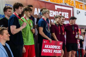 Grupa młodych mężczyzn stojących na podium podczas ceremonii sportowej, z medalami na czerwonych i zielonych wstążkach. Większość ma jasną karnację, krótkie fryzury; jeden z przodu ma jasne, kręcone blond włosy. Ubrani są w stroje sportowe: koszulki granatowe, bordowe oraz jedna zielona w jasny wzór; część ma krótkie spodenki. Kilku zawodników uśmiecha się i obejmuje się ramionami. Z przodu trzymany jest duży czerwony plakat z białym napisem: „AKADEMICKI MISTRZ POLSKI”. W tle widać duży baner z częściowo widocznym napisem.