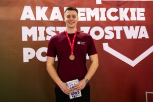 Mężczyzna z medalem na czerwonej wstążce stojący na tle czerwono-brązowego baneru z dużym białym napisem. Mężczyzna ma jasną karnację, krótkie brązowe włosy i uśmiecha się. Ma na sobie bordową koszulkę z małym białym logo i napisem „Akademicki Związek Sportowy” po lewej stronie klatki piersiowej. Na szyi wisi okrągły medal w kolorze brązowym lub miedzianym. Na lewym nadgarstku jest zegarek z metalową bransoletą. W dłoniach trzyma niewielki przedmiot przypominający książeczkę lub folder.