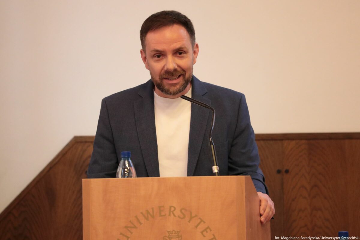 Mężczyzna w średnim wieku stojący za mównicą. Ma krótkie ciemne włosy, przystrzyżoną brodę i jasną karnację. Ubrany jest w biały podkoszulek i granatową marynarkę. Przed nim znajduje się drewniana mównica z okrągłym herbem i napisem „UNIWERSYTET” w dolnej części, a na górze stoi mała plastikowa butelka wody z niebieską zakrętką. Z mównicy wystaje cienki, czarny mikrofon ustawiony na wysokość jego ust. Tło jest jednolite, w jasnym kolorze, dolna część ściany wykończona jest ciemnym drewnem. W prawym dolnym rogu zdjęcia małym drukiem widoczny jest podpis: „fot. Magdalena Seredyńska/Uniwersytet Szczeciński”.