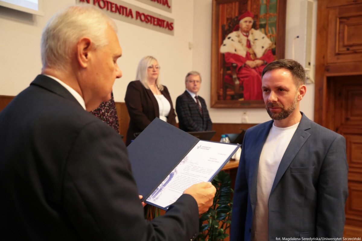 Uroczyste wręczenie dokumentu w sali uczelni. Na pierwszym planie po lewej stronie starszy mężczyzna o siwych włosach w ciemnym garniturze trzyma otwartą niebieską teczkę z dokumentem i czyta go na głos w stronę mężczyzny stojącego naprzeciwko. Po prawej stronie stoi młodszy mężczyzna o jasnej karnacji, krótkich ciemnych włosach i przystrzyżonej brodzie, ubrany w jasny T‑shirt i niebieską marynarkę; patrzy uważnie na osobę z dokumentem, ma poważny wyraz twarzy. W tle za nimi stoją dwie osoby: kobieta o jasnych włosach do ramion w okularach, w białej bluzce i ciemnej marynarce, oraz mężczyzna w okularach w kraciastej marynarce i koszuli z krawatem; oboje wyglądają, jakby uczestniczyli w oficjalnym posiedzeniu. Na ścianie z tyłu widoczny jest duży obraz przedstawiający siedzącego dostojnika akademickiego w czerwonym stroju z białą peleryną z czarnymi elementami. Po lewej stronie, wysoko na ścianie, znajduje się fragment napisu po łacinie w czerwonych literach: „IN POTENTIA POSTENTAS” (część tekstu jest ucięta kadrem). W pomieszczeniu dominują drewniane elementy wystroju, w tym drzwi po prawej stronie; na stole w tle stoją laptopy i butelki z wodą, co podkreśla charakter oficjalnego, akademickiego wydarzenia.