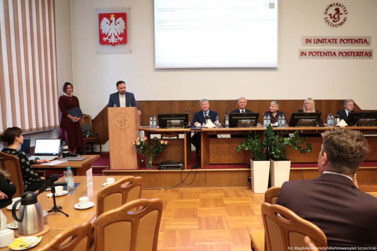 Sala Senatu US na uniwersytecie podczas oficjalnego zebrania. Na środku kadru stoi mężczyzna przy jasnej drewnianej mównicy z logo Uniwersytetu Szczecińskiego; mówi do mikrofonu. Obok niego, po lewej stronie, stoi kobieta w bordowej, dość długiej sukience, trzyma teczkę lub segregator. Za mównicą ciągnie się długi stół prezydialny; przy nim siedzi sześć osób w eleganckich strojach, głównie w garniturach i marynarkach. Przed każdym z nich stoi monitor, butelki wody i mikrofony. Za stołem, w tle, znajduje się duży ekran z wyświetlonym tekstem oraz herb Polski z białym orłem na czerwonym tle. Na prawej ścianie widnieje napis po łacinie „IN UNITATE POTENTIA, IN POTENTIA POSTERITAS” oraz okrągłe logo z napisem „UNIWERSYTET SZCZECIŃSKI”. Na pierwszym planie stoi kilka stołów konferencyjnych z drewnianymi krzesłami; na stołach widać filiżanki, dzbanki z napojami, mikrofony i laptopy. Pod stołem prezydialnym stoją dwie większe donice z zielonymi roślinami oraz bukiet róż przy mównicy. Podłoga jest z jasnego parkietu, a po lewej stronie sali widać wysokie, pionowe żaluzje okienne. W dolnym prawym rogu znajduje się mały napis z informacją o autorce zdjęcia i uczelni.