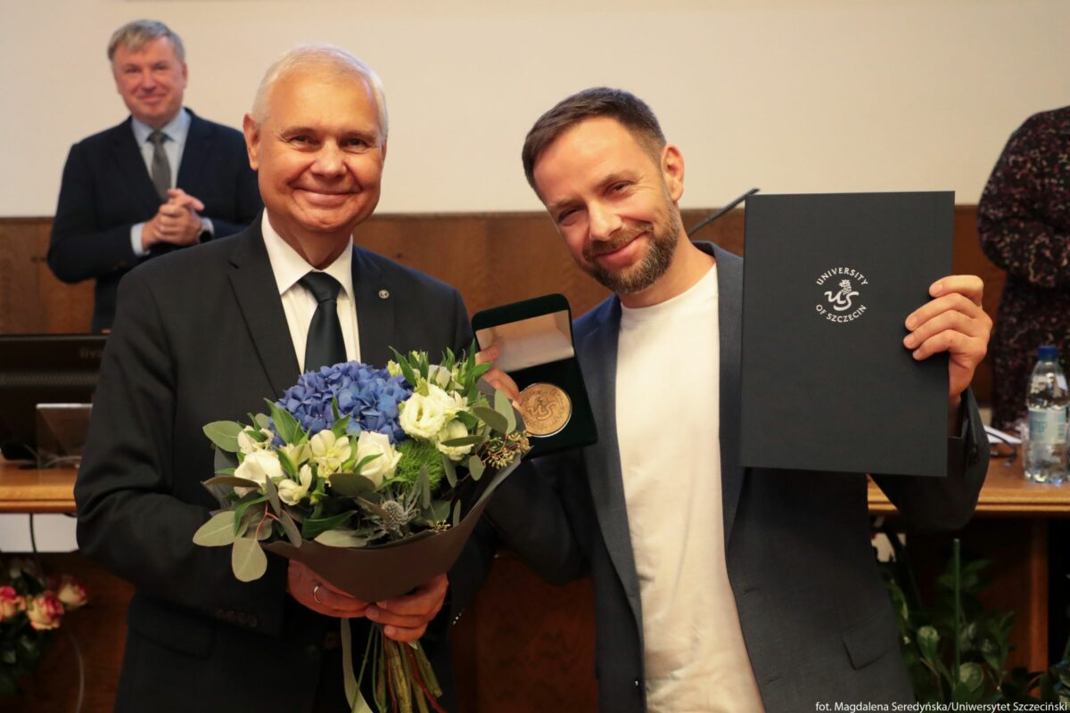 Dwóch mężczyzn stojących obok siebie w sali uroczystej. Po lewej stronie znajduje się starszy mężczyzna o jasnych włosach i jasnej karnacji, ubrany w ciemny garnitur, białą koszulę i ciemny krawat. Uśmiecha się i trzyma przed sobą duży bukiet świeżych kwiatów w papierowej osłonce; w bukiecie są głównie niebieskie hortensje oraz białe kwiaty z zielonymi liśćmi. Po prawej stronie stoi młodszy mężczyzna o jasnej karnacji, krótkich ciemnych włosach i brodzie, ubrany w biały t‑shirt i ciemną marynarkę. Również się uśmiecha. W jednej dłoni trzyma otwarte pudełko z okrągłym medalem w środku, w drugiej ciemną teczkę lub dyplom z logo i napisem w języku angielskim „University of Szczecin” oraz graficznym znakiem przypominającym stylizowaną literę S lub węża. W tle widać rozmazaną postać mężczyzny w garniturze, który stoi przy biurku lub mównicy i ma złożone dłonie, jakby klaskał. Za nim znajduje się drewniany panel ścienny oraz sprzęt biurowy. Po prawej stronie w tyle stoi kolejna osoba i stoi butelka wody na stole. Cała scena sprawia wrażenie ceremonii nagrodowej lub akademickiego wyróżnienia.