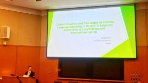 Sala wykładowa. W lewym dolnym rogu mównica i wykładowczyni. Na środku od góry ekran z tytułem prezentacji w języku angielskim (w tłumaczeniu: „Aktualna sytuacja i wyzwania edukacji chińskiej w Polsce”).