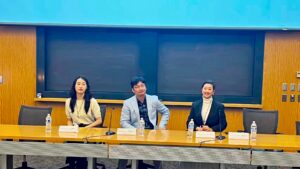 Sala wykładowa. Za stołem siedzi troje prelegentów (2 kobiety i mężczyzna w środku). Na stole butelki z wodą oraz imiona i nazwiska wykładowców.