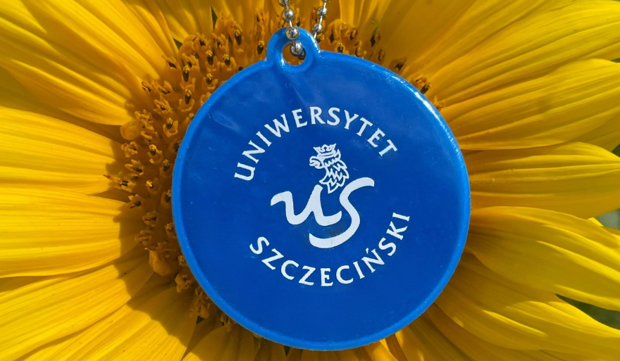 Niebieski, okrągły odblask z białym logiem Uniwersytetu Szczecińskiego na tle żółtego słonecznika.
