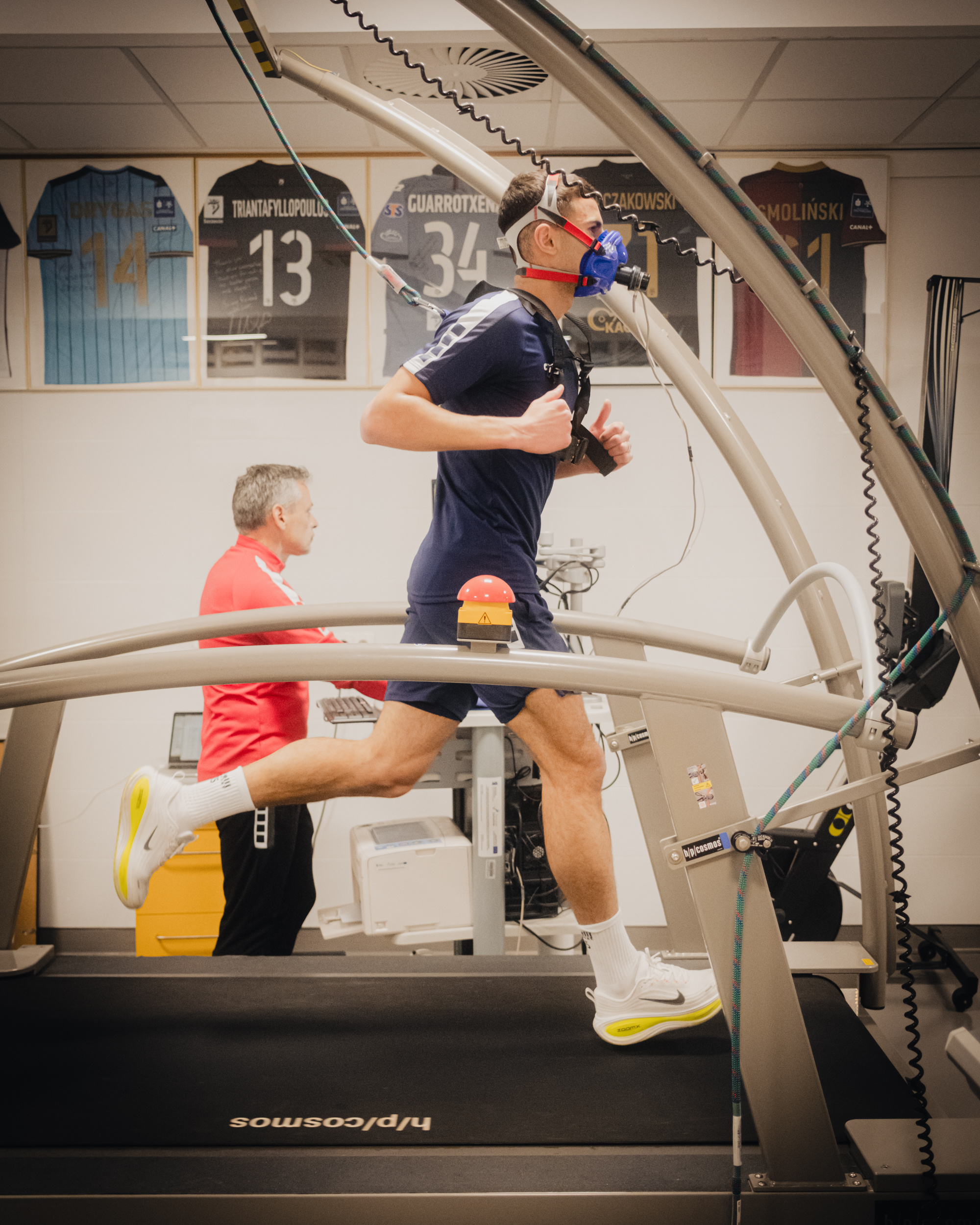 Mężczyzna biegnący na bieżni w laboratorium sportowym, z maską do pomiaru oddechu i uprzężą z przewodami. Ma jasną karnację, krótkie ciemne włosy, granatowy strój sportowy, białe skarpety i białe buty z żółtymi elementami. Bieżnia jest duża i otoczona metalową konstrukcją z kablami; na poręczy znajduje się czerwony przycisk awaryjny. W tle siedzi starszy mężczyzna o siwych włosach w czerwonej bluzie, obok sprzętu komputerowego. Na ścianie wiszą oprawione koszulki sportowe z numerami i nazwiskami.