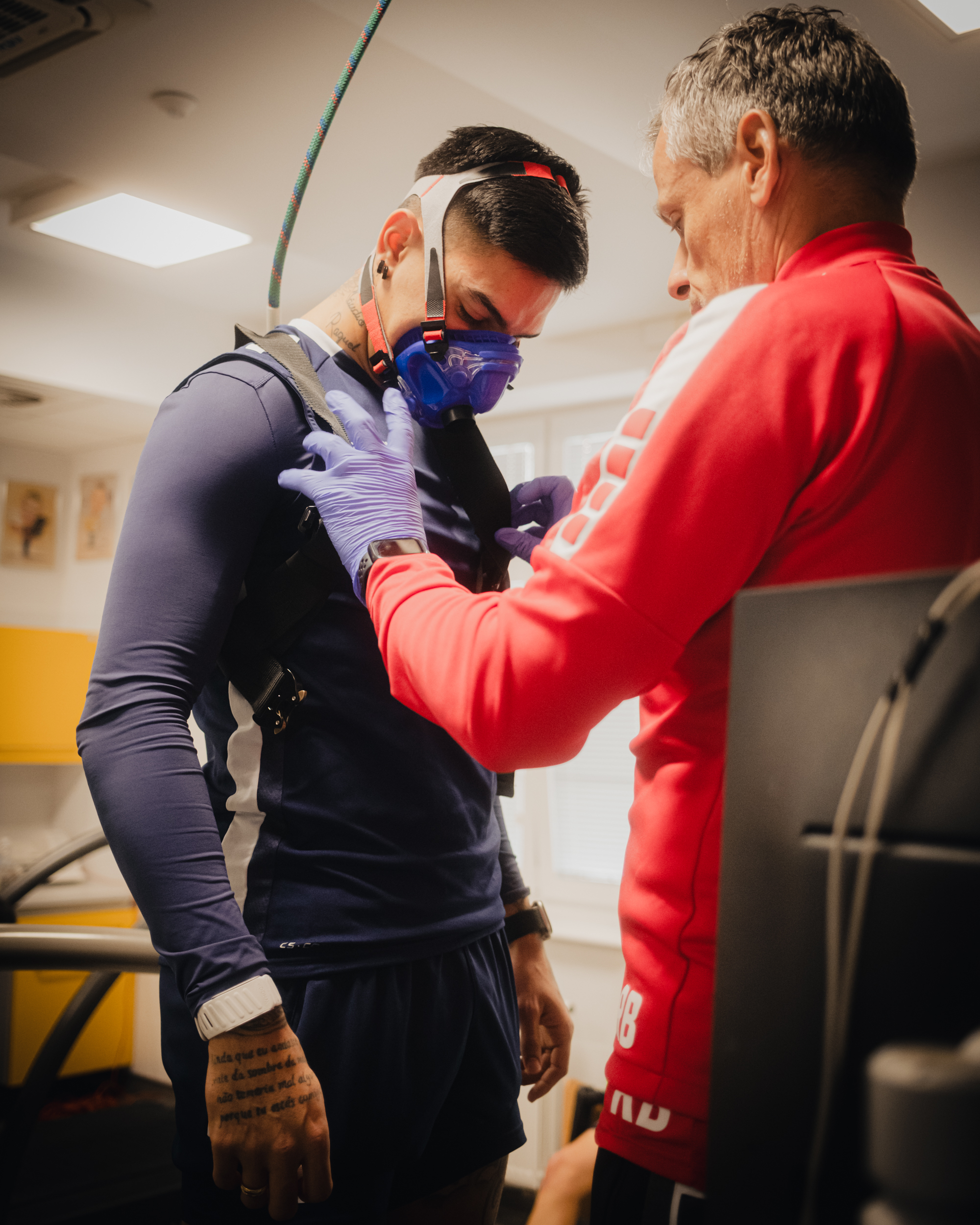 Dwóch mężczyzn w pomieszczeniu treningowo-medycznym, jeden w masce oddechowej i uprzęży z przewodem, drugi w czerwonej bluzie i rękawiczkach jednorazowych, regulujący sprzęt na klatce piersiowej. Mężczyzna po lewej ma krótkie, ciemne włosy i średnią karnację. Ma na sobie granatową, obcisłą koszulkę z długim rękawem i granatowe spodenki. Na twarzy ma niebieską maskę oddechową z paskami na głowie, a na ramionach i klatce piersiowej czarną uprząż; do maski prowadzi gruby, elastyczny przewód idący w górę kadru. Na nadgarstku ma białą opaskę/zegarek sportowy, a na dłoni i szyi widać tatuaże. Głowa jest pochylona w dół, jakby skupiał się na zakładanym sprzęcie. Mężczyzna po prawej ma krótkie, siwiejące włosy i jasną karnację. Jest pokazany z profilu, ubrany w czerwoną bluzę z białymi elementami i ma na rękach niebiesko-fioletowe rękawiczki. Rękami dopina lub poprawia elementy uprzęży na klatce piersiowej drugiego mężczyzny. W tle widać sufit z jasnymi panelami oświetleniowymi oraz fragmenty sprzętu/urządzeń, co sugeruje gabinet testów wysiłkowych lub salę treningową.