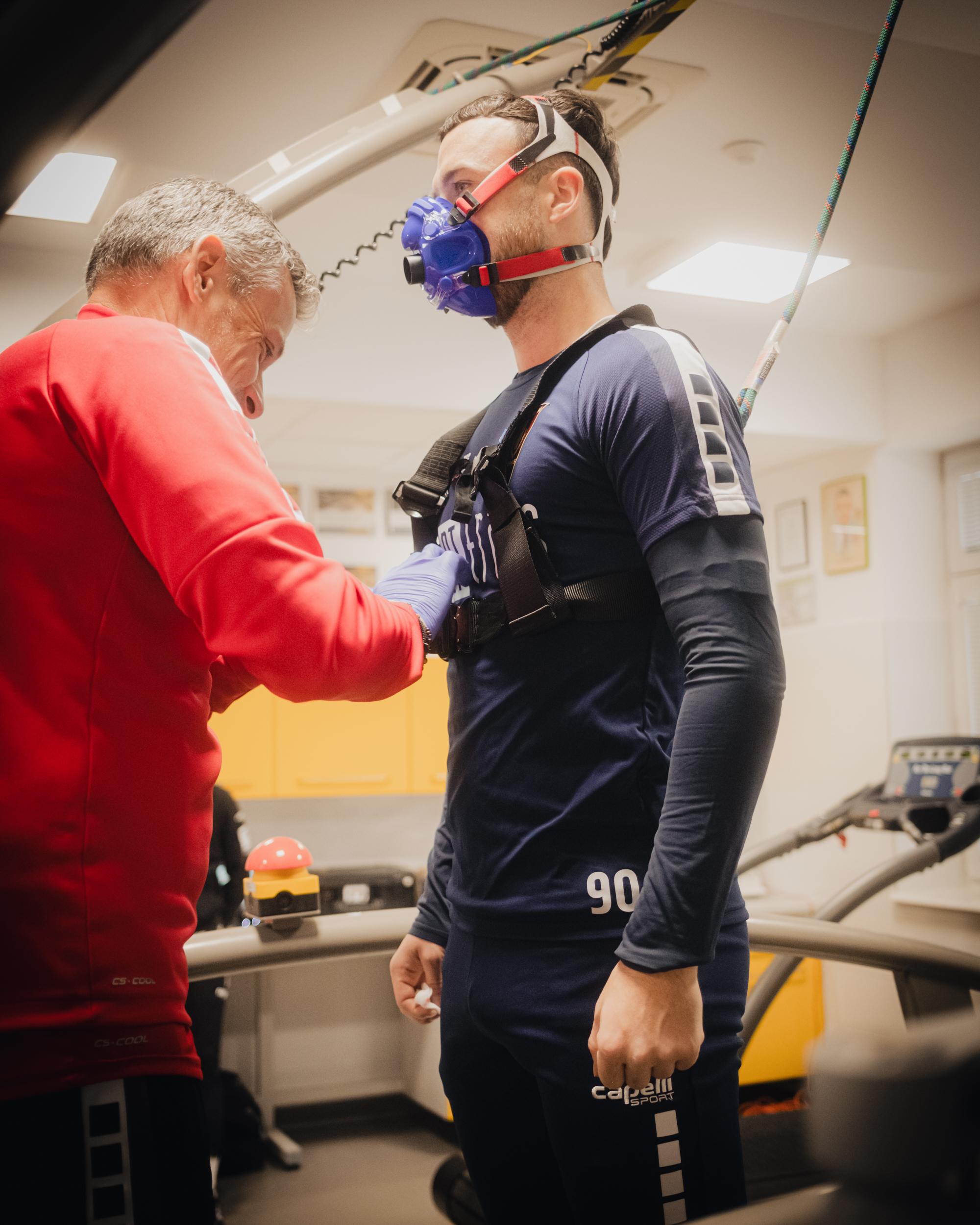 Dwóch mężczyzn w laboratorium wysiłkowym, podczas testu wydolnościowego. Mężczyzna po prawej stronie stoi w profilu i ma na twarzy niebieską maskę oddechową z czerwono-czarnymi paskami mocującymi na głowie. Jest w granatowym, sportowym stroju z długim rękawem i dopasowanych spodniach; na udzie widoczny jest biały napis „capelli sport”, a na biodrze liczba „90”. Na klatce piersiowej ma czarną uprząż/pas z klamrami. Mężczyzna po lewej stronie, w czerwonej bluzie, pochyla się i zakłada lub reguluje elementy przy pasie/uprzęży na klatce piersiowej drugiego mężczyzny; ma siwe włosy i rękawiczkę w jasnofioletowym kolorze. W tle widać sprzęt do ćwiczeń (bieżnia) i pomieszczenie z jasnym sufitem oraz oświetleniem.