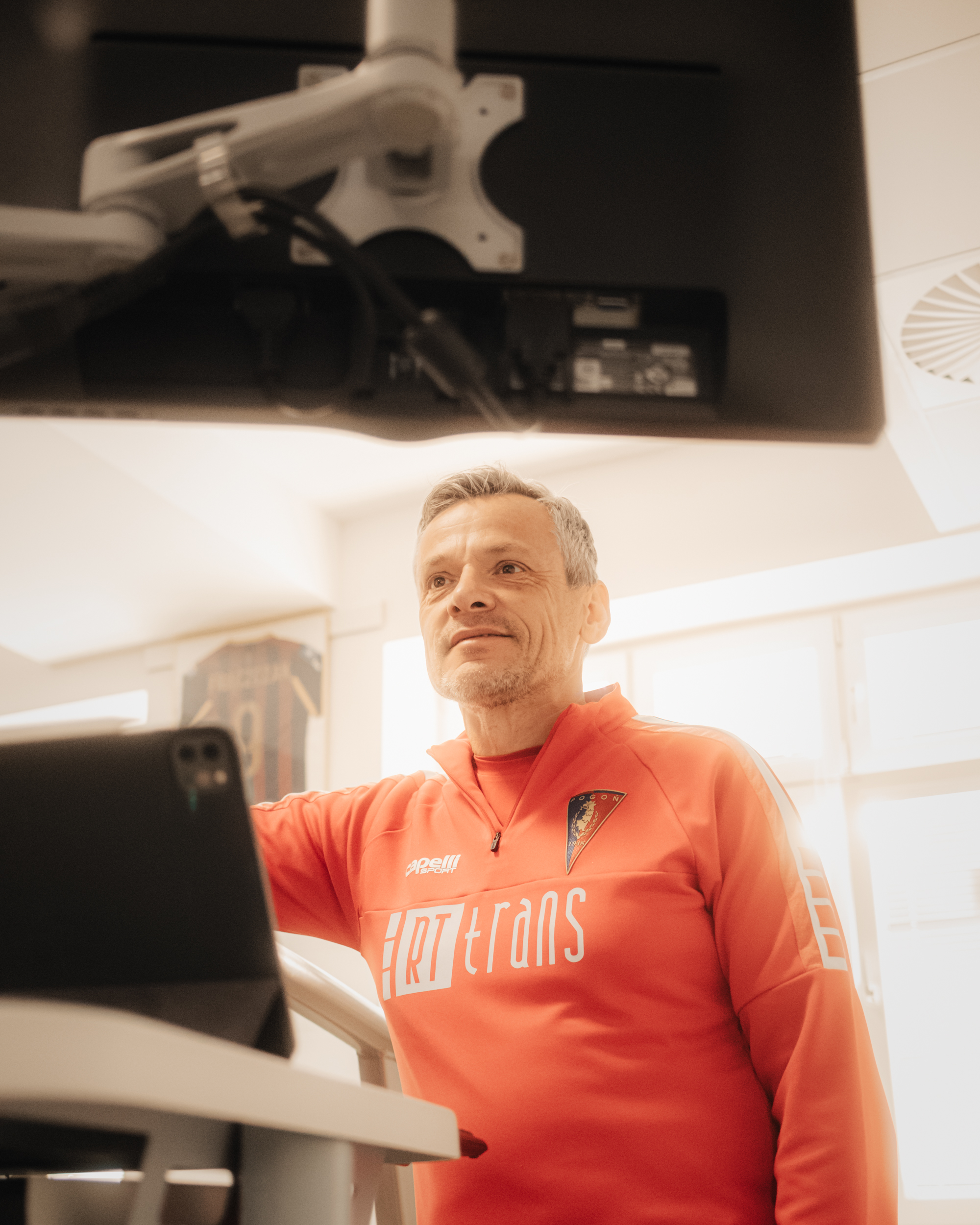 Mężczyzna w pomarańczowej sportowej bluzie stojący przy urządzeniu treningowym. Ma krótkie, siwe włosy i jasną skórę, a na twarzy lekki zarost. Na bluzie widać herb oraz duży napis „HRTtrans”; obok jest też mniejsze logo „capelli”. Ujęcie wygląda na wykonane w jasnym pomieszczeniu, a u góry kadru widać spód dużego monitora na ramieniu.