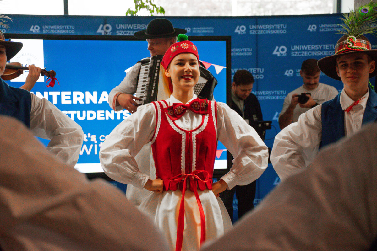 Grupa osób na scenie podczas występu folklorystycznego, z kobietą na pierwszym planie w stroju ludowym. Kobieta stoi na środku z rękami na biodrach i uśmiecha się. Ma jasną cerę, czerwony gorset z białymi zdobieniami na białej bluzce z bufiastymi rękawami oraz białą spódnicę; w pasie zawiązana jest długa czerwona wstążka. Na głowie ma czerwone nakrycie z zielonym pomponem i haftowanymi kwiatami, a przy szyi dużą czerwoną kokardę z wzorem kwiatowym. Po lewej stronie widać fragment muzyka grającego na skrzypcach. Z tyłu, za kobietą, stoi mężczyzna w kapeluszu i gra na akordeonie (na pasku widać napis „WELTMEISTER”). Po prawej stronie jest mężczyzna w białej koszuli i niebieskiej kamizelce, w kapeluszu ozdobionym piórami. W tle znajduje się niebieska ścianka z powtarzającym się napisem „40 LAT UNIWERSYTETU SZCZECIŃSKIEGO”. Obok jest duży ekran z częściowo widocznym tekstem „INTERNATIONAL STUDENTS”.