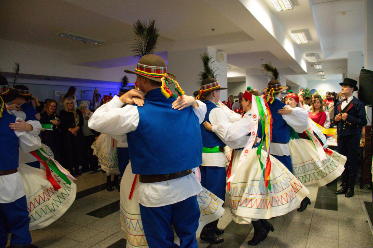 Grupa tancerzy w strojach ludowych tańczy w sali wewnątrz budynku. Mężczyźni mają białe koszule, niebieskie kamizelki i spodnie oraz brązowe kapelusze z piórami i kolorowymi wstążkami. Kobiety mają białe, rozkloszowane spódnice z kolorowymi wzorami przy dole oraz długie, wielobarwne wstążki przy strojach i na głowach. Pary trzymają się w tańcu, a spódnice są rozchylone ruchem. W tle stoi publiczność, a po prawej stronie widoczny jest mężczyzna w ciemnym stroju z cylindrem. Oświetlenie jest sufitowe, a w tle widać niebieskawe światło.