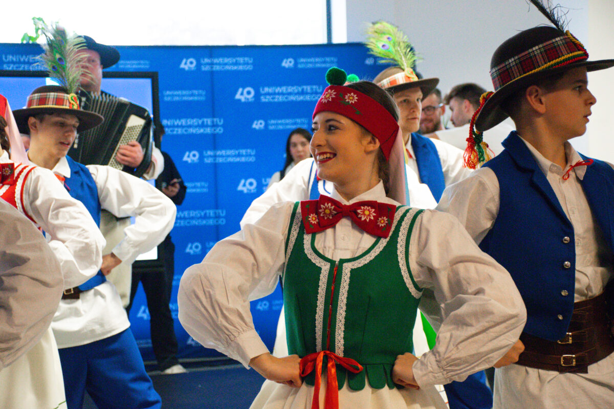 Grupa młodych tancerzy w strojach ludowych podczas występu w pomieszczeniu. Na pierwszym planie pośrodku jest młoda kobieta o jasnej karnacji, uśmiechnięta, z rękami opartymi na biodrach. Ma czerwoną opaskę na głowie z białymi wzorami kwiatowymi, białą bluzkę oraz zielony gorset/sukienkę z białą lamówką i czerwono-zielonymi akcentami. Pod szyją ma dużą czerwoną kokardę z białymi motywami kwiatowymi, a w pasie czerwono-żółtą wstążkę. Wokół niej stoją młodzi mężczyźni w białych koszulach i niebieskich kamizelkach oraz spodniach. Na głowach mają ciemne kapelusze z kolorowymi, kraciastymi opaskami, piórami i ozdobnymi chwostami. W tle widać mężczyznę grającego na akordeonie oraz niebieską ściankę z powtarzającym się napisem „UNIWERSYTETU SZCZECIŃSKIEGO” i liczbą „40”.