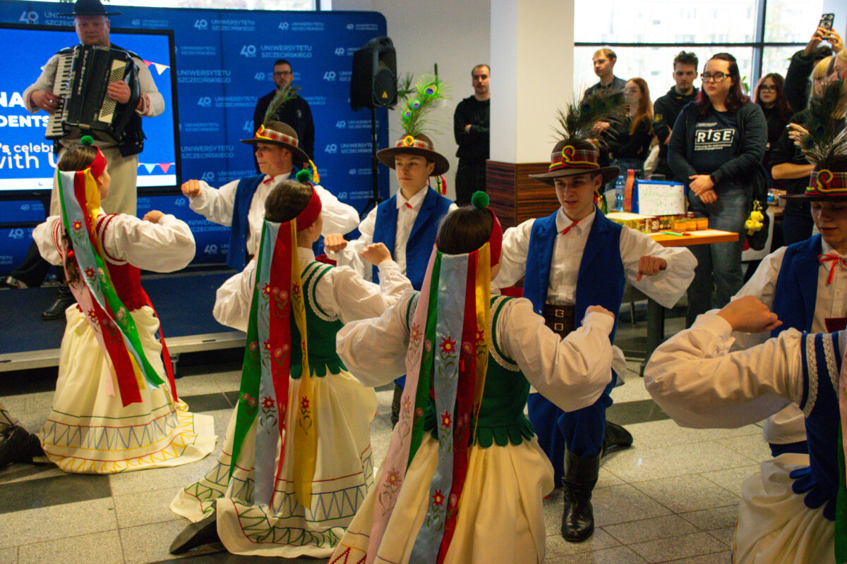 Grupa tancerzy w strojach ludowych podczas występu w pomieszczeniu, z muzykiem grającym na akordeonie na podwyższeniu w tle. W centrum kilka par tańczy w kręgu, z uniesionymi i zgiętymi w łokciach rękami. Tancerze mają jasną karnację. Kobiety są w białych bluzkach, zielonych gorsetach i jasnych, szerokich spódnicach z kolorowymi zdobieniami przy dole. Z tyłu głowy mają długie, wielokolorowe wstążki zwisające na plecy, część z haftowanymi kwiatami. Mężczyźni są w białych koszulach, niektórzy w niebieskich kamizelkach i spodniach, z czerwonymi wiązaniami pod szyją. Na głowach mają kapelusze z piórami (wyglądają jak pawie). W tle stoi publiczność przy ścianie i obok stołu z różnymi przedmiotami. Za muzykiem jest niebieska ścianka z powtarzającym się napisem „UNIWERSYTETU SZCZECIŃSKIEGO” oraz „40”.
