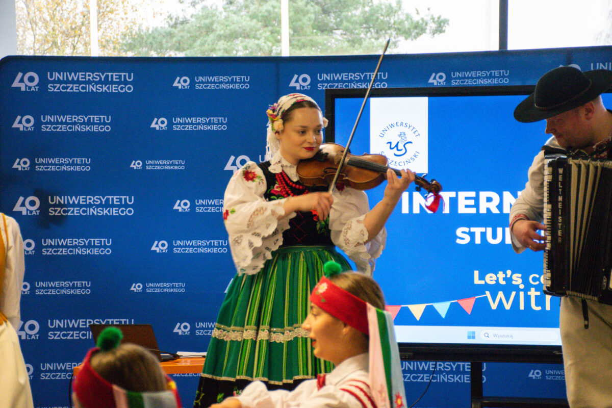 Występ muzyczny na tle ścianki z napisem „40 LAT UNIWERSYTETU SZCZECIŃSKIEGO”. Na środku stoi kobieta o jasnej skórze w stroju ludowym, grająca na skrzypcach; ma białą haftowaną bluzkę, czarny gorset z kolorowymi kwiatami i zieloną spódnicę w pionowe pasy, a na głowie chustę. Po prawej stronie częściowo widać mężczyznę o jasnej skórze w kapeluszu, grającego na akordeonie; na instrumencie widoczny jest napis „WELTMEISTER”. W dolnej części kadru widać fragmenty osób w strojach ludowych, z czerwonymi opaskami na głowie. Z tyłu jest duży ekran z logo „Uniwersytet Szczeciński” .