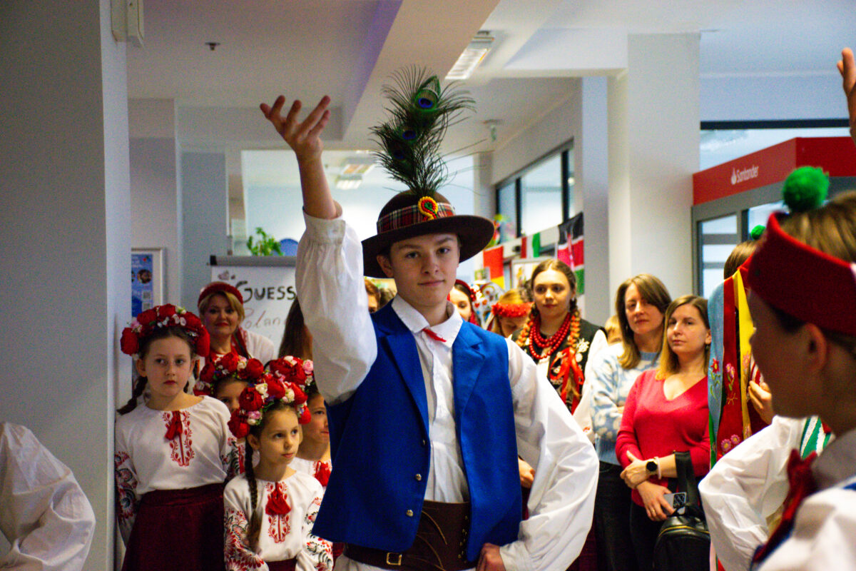 Młody chłopak w stroju ludowym stojący na korytarzu, z uniesioną do góry prawą ręką i lewą dłonią opartą na biodrze. Ma na sobie białą koszulę z szerokimi rękawami, niebieską kamizelkę i ciemny pas. Na głowie ma kapelusz z wysokim piórem pawia. W tle stoi grupa dzieci i dorosłych, część w tradycyjnych strojach: dzieci w białych haftowanych bluzkach i wiankach z czerwonych kwiatów, a kobiety m.in. w czerwonych koralach i wzorzystych elementach ubioru. Po prawej stronie kadru widać fragment osoby w podobnym stroju ludowym z czerwonym nakryciem głowy. Scena wygląda na wydarzenie w budynku, na jasnym korytarzu z flagami i dekoracjami w tle.