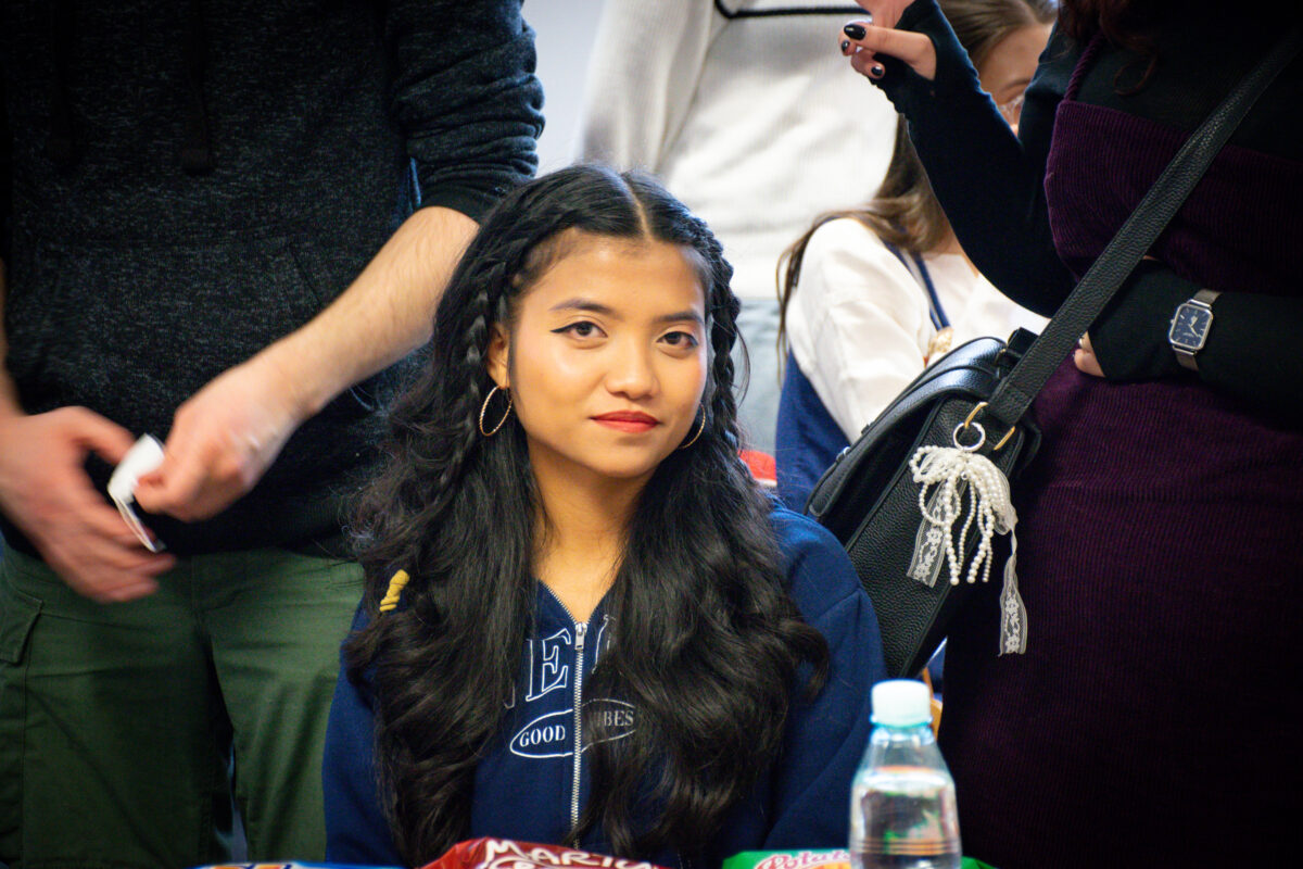 Młoda kobieta siedząca na pierwszym planie, otoczona przez stojących ludzi. Ma długie, czarne, falowane włosy z dwoma warkoczami przy przedziałku, jasnobrązową cerę, delikatny makijaż z wyraźną kreską na powiekach i czerwone usta. Ma duże kolczyki-koła i granatową bluzę z zamkiem oraz białymi napisami. Wokół niej widać tułowie kilku osób: po lewej mężczyznę w ciemnym swetrze i zielonych spodniach, po prawej osobę w czarno-fioletowej sukience z paskiem i zegarkiem, z czarną torbą przewieszoną przez ramię (zawieszona biała ozdoba z koralików/wstążek). Na dole kadru widać fragment stołu z plastikową butelką wody i częścią opakowań od przekąsek.