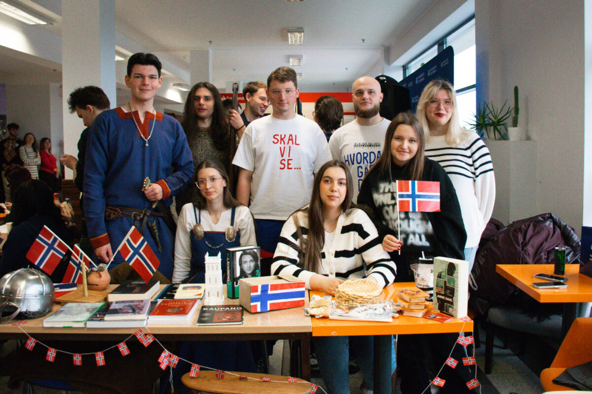 Grupa młodych osób stojących i siedzących za stołem z norweskimi flagami i przekąskami w jasnym pomieszczeniu, prawdopodobnie na szkolnym lub uczelnianym wydarzeniu. Na pierwszym planie stoi długi stół udekorowany girlandą z małych norweskich flag. Na blacie leżą książki, broszury i kilka przedmiotów związanych z Norwegią; widać też metalowy hełm wikinga po lewej stronie. Na środku i po prawej stronie są wafle i inne jedzenie na talerzach oraz w folii, a także pudełka i kubki. Na stole i w rękach kilku osób są większe norweskie chorągiewki na patyczkach. Za stołem jest około dziewięciu osób. Część siedzi, część stoi. Ubrania są codzienne; jedna osoba po lewej ma niebieski strój stylizowany na historyczny, z pasem. W centrum stoi osoba w białej koszulce z czerwonym napisem „SKAL VI SE…”. Po prawej stronie stoi osoba w białym swetrze w czarne poziome paski i ma jasne włosy oraz okulary. W tle widać innych uczestników wydarzenia i elementy sali z dużymi oknami po prawej.