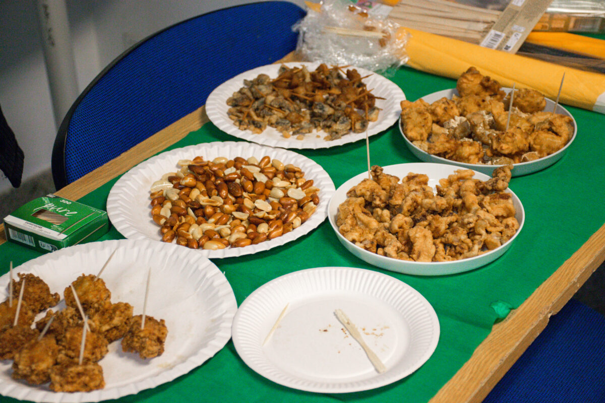 Stół przykryty zielonym obrusem z kilkoma jednorazowymi papierowymi talerzami i miskami z przekąskami. Na stole są porcje orzechów (mieszanka jasnych i brązowych) oraz kilka naczyń z panierowanymi, smażonymi kawałkami jedzenia; część kawałków ma włożone wykałaczki. Z przodu leży pusty papierowy talerz z pojedynczą wykałaczką i okruszkami. Po lewej stronie stoi małe zielone pudełko. W tle widać niebieskie krzesła oraz opakowania i materiały leżące na blacie.