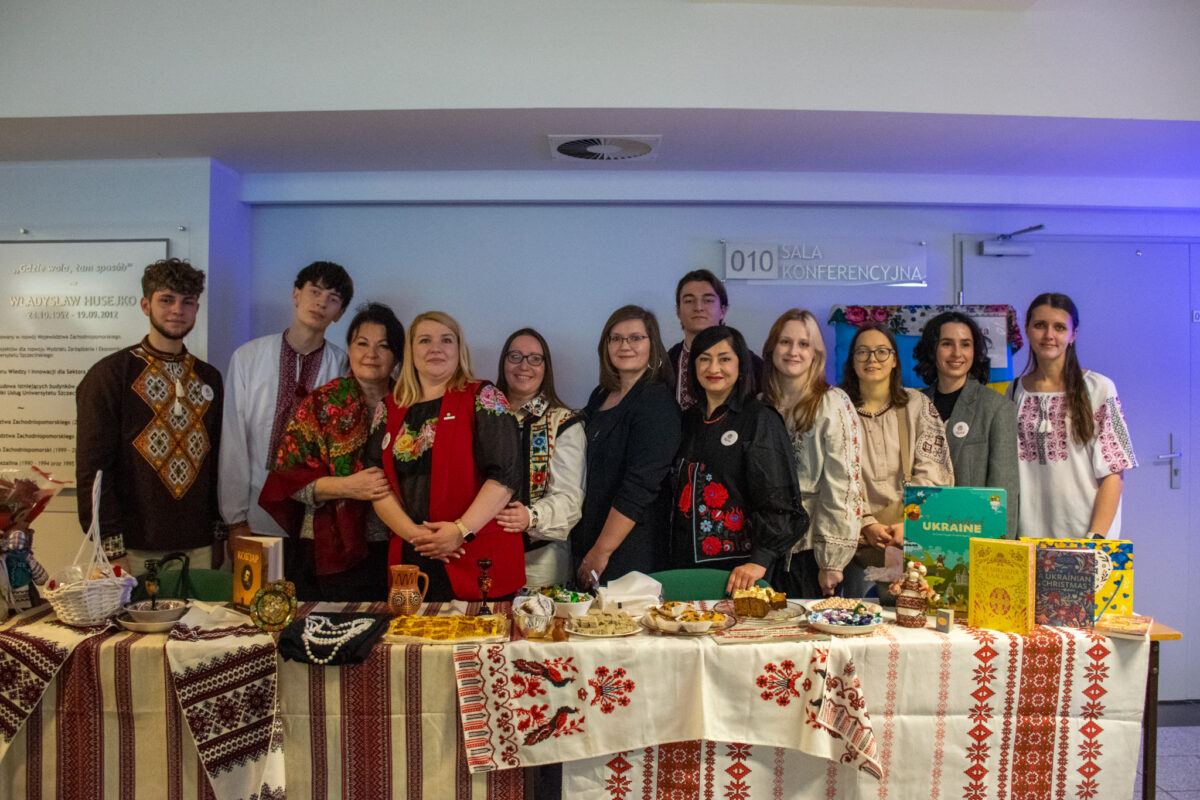Grupa kilkunastu osób stojąca za długim stołem nakrytym haftowanymi, ludowymi obrusami. Osoby pozują do wspólnego zdjęcia w pomieszczeniu opisanym tabliczką „010 SALA KONFERENCYJNA”; część ma na sobie ubrania z tradycyjnymi, haftowanymi wzorami w stylu ukraińskim. Na stole leżą i stoją różne przedmioty oraz poczęstunek: miski, talerze z jedzeniem, ozdoby i rękodzieło, a także kilka książek; po prawej stronie wyraźnie widoczna jest książka z tytułem „UKRAINE”. W tle po lewej wisi duża tablica z dłuższym tekstem (nieczytelnym w całości na tym ujęciu), a po prawej są drzwi i fragmenty kolorowych dekoracji.