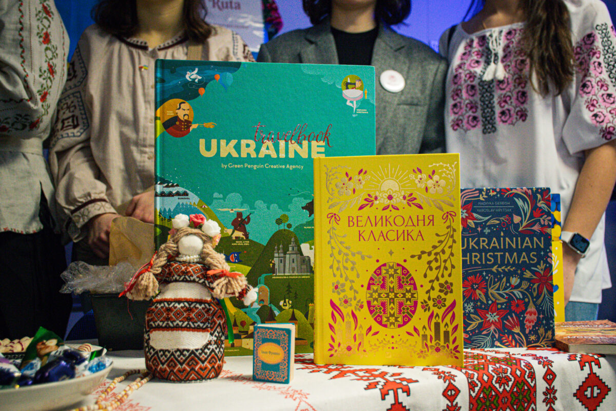 Stół z wystawionymi książkami i ozdobami w ukraińskim stylu, a w tle stoją trzy osoby w haftowanych bluzkach i marynarce. Na środku stoi duża książka z okładką w odcieniach turkusu i zieleni z napisem „travelbook UKRAINE” oraz „by Green Penguin Creative Agency”. Przed nią stoi mała lalka-motanka w biało-czerwono-czarnym wzorzystym stroju, z warkoczami i czerwonymi wstążkami. Po prawej stronie jest żółta książka z ukraińskim tytułem „ВЕЛИКОДНЯ КЛАСИКА”, z dekoracyjnymi ornamentami i dużym krzyżem pośrodku. Jeszcze dalej po prawej stoi granatowa książka z napisem „UKRAINIAN CHRISTMAS” wśród czerwono-niebieskich kwiatowych wzorów. Na stole leży biały obrus z czerwono-czarnym haftowanym wzorem. Z lewej strony widać miskę z zawiniętymi cukierkami oraz inne drobne przedmioty, a obok stoi ciemny pojemnik lub koszyk. W tle na górze widać fragment napisu, w którym czytelne jest słowo „Ruta”.