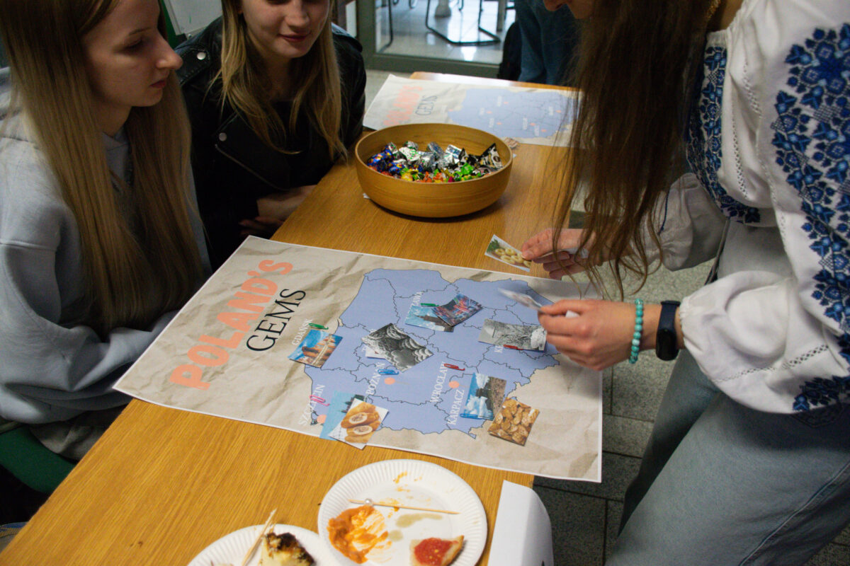 Grupa młodych osób siedzących przy drewnianym stole nad dużą planszą z mapą Polski. Na planszy widać napis „POLAND’S GEMS” oraz mapę z zaznaczonymi regionami i naklejonymi obrazkami (m.in. jedzenie i krajobrazy). Jedna osoba w białej bluzie z niebieskim haftem pochyla się nad mapą i układa lub przykleja kolejny obrazek. Na stole stoi miska z cukierkami w papierkach. Na dole kadru są papierowe talerzyki z resztkami jedzenia i sosu.