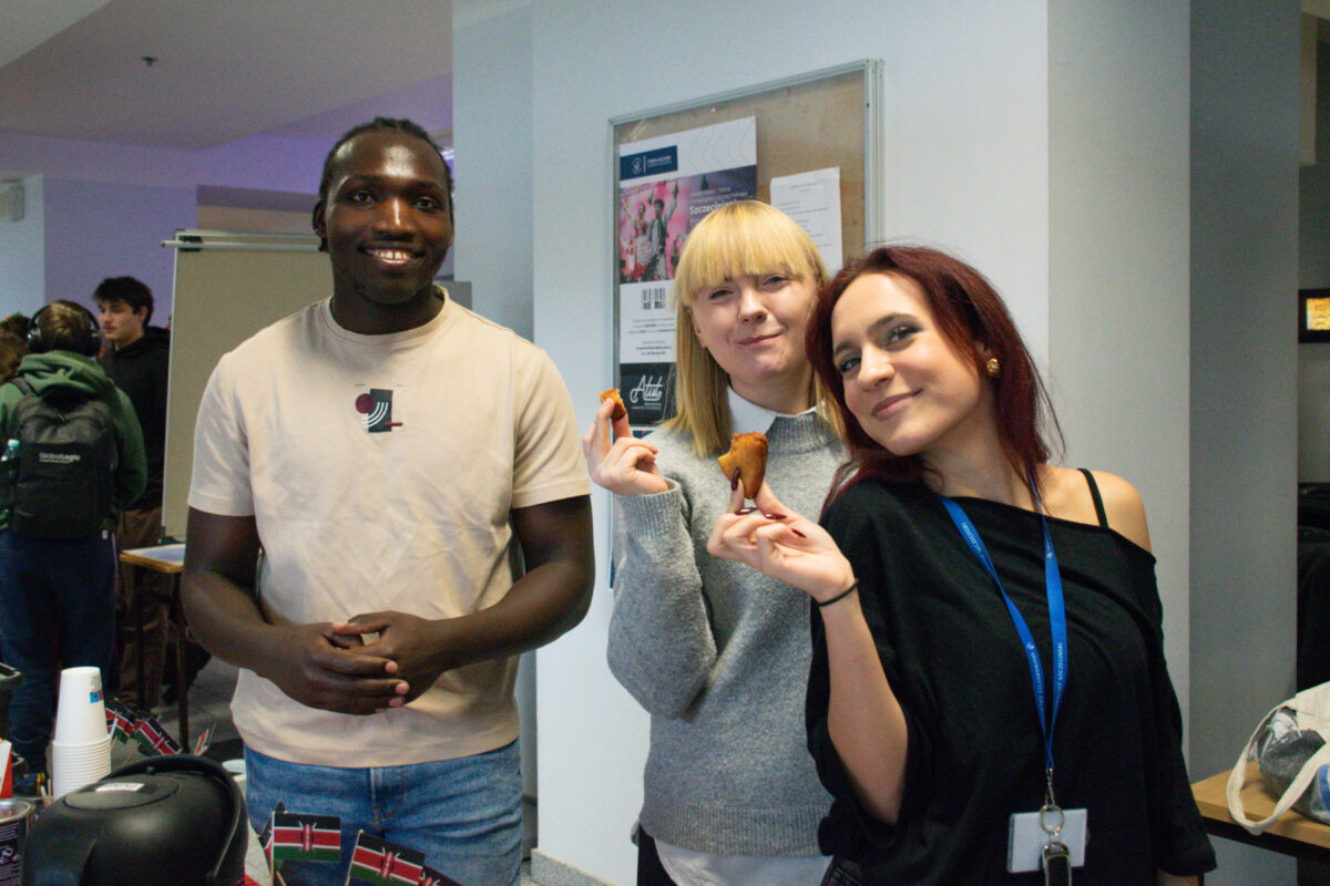 Grupa trzech osób stojących w pomieszczeniu, ujętych od pasa w górę. Po lewej stoi mężczyzna o ciemnej skórze i krótkich, zaplecionych włosach, uśmiecha się i ma na sobie jasny T-shirt oraz niebieskie dżinsy; dłonie ma złożone przed sobą. Po prawej stoją dwie kobiety blisko siebie; obie trzymają w dłoniach małe, brązowe przekąski. Kobieta z tyłu ma jasną skórę i proste blond włosy z grzywką, ubrana jest w szary sweter. Kobieta z przodu ma jasną skórę i długie, ciemnoczerwone włosy, ubrana jest w czarną bluzkę odsłaniającą jedno ramię i ma na szyi niebieską smycz z identyfikatorem. W tle widać tablicę ogłoszeń na ścianie oraz kilka osób i elementy stołu po lewej stronie.