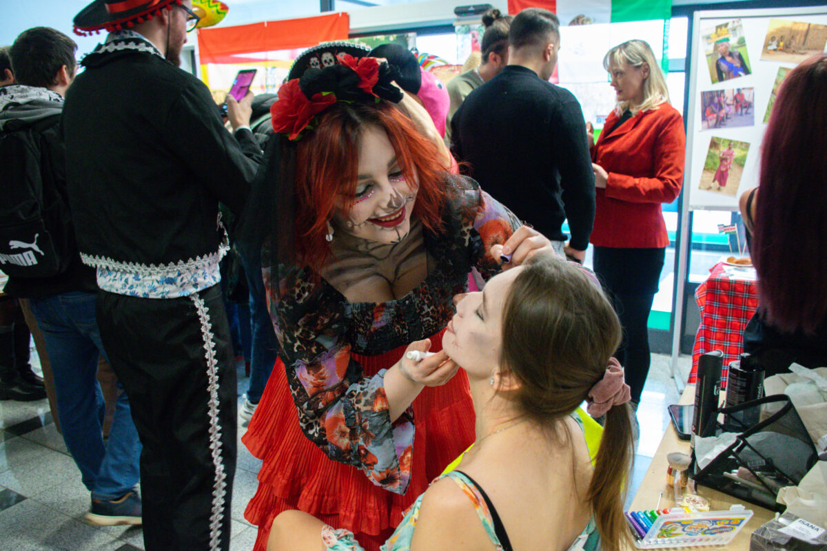 Tłum ludzi na wydarzeniu w pomieszczeniu, z kobietą w czerwonej spódnicy i w czarnym nakryciu głowy z czerwonymi kwiatami, która wykonuje makijaż siedzącej kobiecie. Na pierwszym planie osoba z rudymi włosami, z makijażem w stylu „dzień zmarłych” (biała twarz, czarne i czerwone detale przy oczach i ustach), pochyla się nad kobietą z jasną skórą i brązowymi włosami związanymi w kucyk. Siedząca kobieta ma na sobie jasną, wzorzystą sukienkę na ramiączkach; osoba stojąca trzyma przy jej twarzy narzędzie do makijażu, jakby domalowywała szczegół przy policzku lub ustach. Wokół widać dużo osób w strojach tematycznych; po lewej ktoś ma czarny strój z białą koronką i kapelusz w stylu sombrero. W tle są wiszące flagi i tablica/ścianka z kilkoma zdjęciami. Po prawej stoi stolik z kosmetykami i akcesoriami do makijażu oraz telefonem, a obok inny stolik przykryty czerwono‑zieloną kratą.