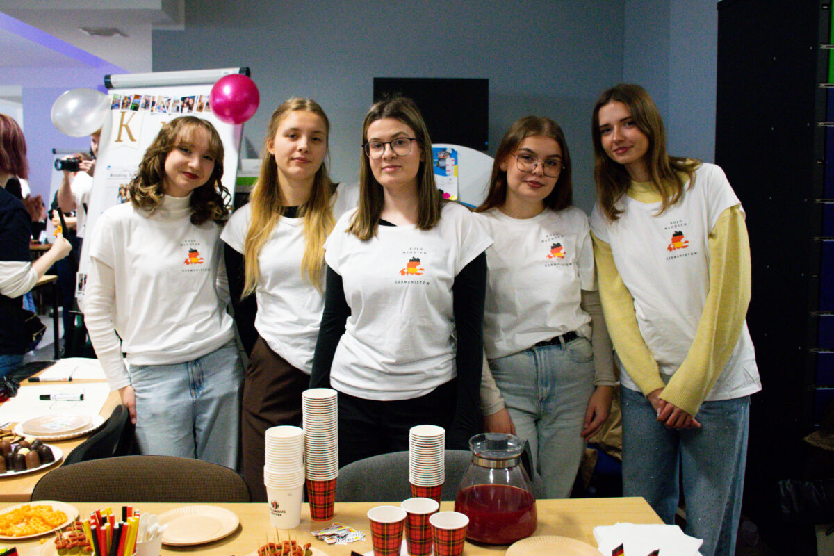 Grupa pięciu młodych kobiet stojących za stołem z przekąskami na imprezie w pomieszczeniu. Mają jasną karnację i pozują do zdjęcia, stojąc blisko siebie. Wszystkie są ubrane w białe koszulki z małym nadrukiem na piersi (widać motyw w barwach czarno-czerwono-żółtych) założone na długi rękaw; mają jeansy lub ciemne spodnie. Dwie osoby mają okulary. Fryzury są różne: od kręconych włosów do długich prostych, w odcieniach brązu i blondu. Na stole stoją wysokie stosy jednorazowych kubków w czerwono-zieloną kratkę oraz biały kubek. Jest też szklany dzbanek z czerwonym napojem, papierowe talerzyki, słomki w pojemniku i kilka misek/talerzy z przekąskami.