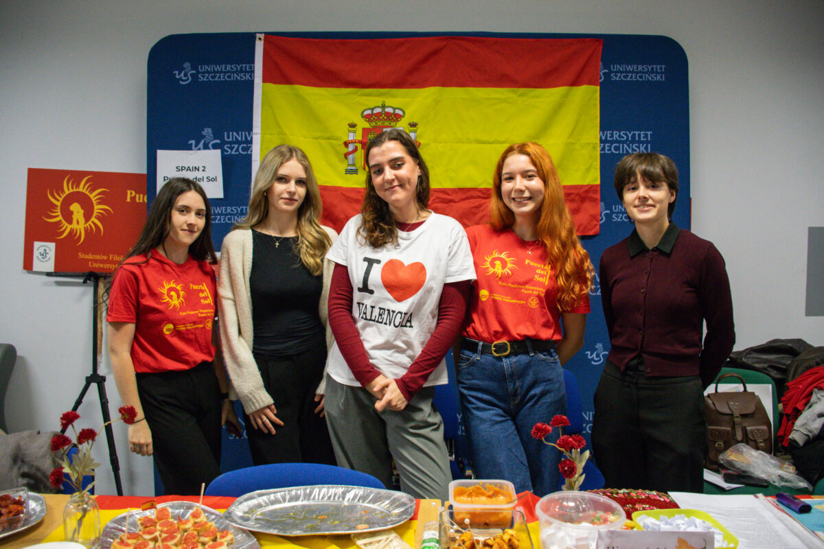 Grupa pięciu osób stojących w pomieszczeniu przed dużą flagą Hiszpanii z herbem pośrodku. Trzy osoby mają na sobie czerwone koszulki z żółtym nadrukiem, jedna osoba biały T-shirt z napisem „I serduszko VALENCIA”, a jedna ciemną koszulę i bordowy sweter. W tle jest niebieska ścianka z powtarzającym się napisem „UNIWERSYTET SZCZECIŃSKI”. Po lewej stronie stoi czerwony plakat z żółtym symbolem słońca i częściowo widocznym tekstem; obok jest kartka z napisem. Z przodu, na stole, stoją tacki i miski z jedzeniem (widać m.in. małe kanapki/koreczki, porcje w pojemnikach), są też małe wazoniki z czerwonymi goździkami. Po prawej na krześle lub blacie leży torba i stos ubrań lub materiałów.