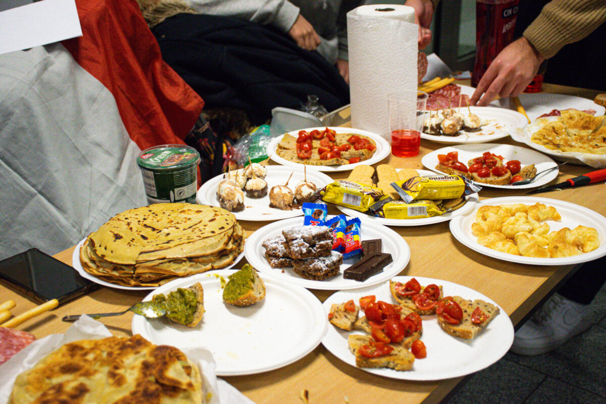 Stół zastawiony jedzeniem na papierowych talerzach. Na środku stoi wysoka rolka ręcznika papierowego. Po lewej stronie leży duży stos naleśników na białym talerzu; obok widać czarny telefon i drewniane pałeczki. Na kilku talerzach są małe kanapki/bruschetty z pokrojonymi czerwonymi pomidorkami. Widać też talerz z małymi kawałkami mięsa lub przekąskami na wykałaczkach. Na jednym talerzu są chipsy. Na innym talerzu leżą żółte, podłużne, paczkowane batoniki; obok są małe niebieskie batony i kilka kostek czekolady. Na talerzu po lewej na dole jest płaski placek lub podpłomyk. W pobliżu ręcznika stoi przezroczysty kubek z czerwonym napojem. W kadrze widać fragmenty rąk kilku osób siedzących przy stole.
