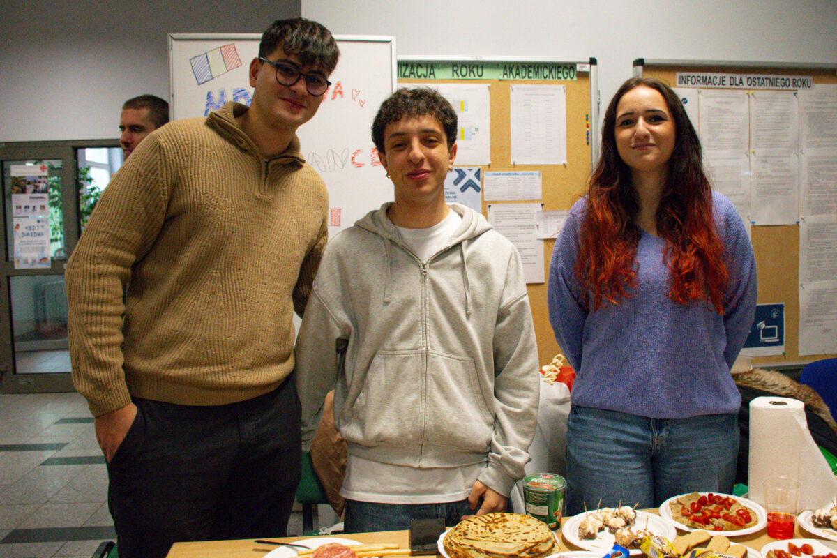 Trzy osoby stojące za stołem z jedzeniem w pomieszczeniu przypominającym szkolny korytarz lub salę. Po lewej stoi młody mężczyzna o jasnej skórze, w okularach, z krótkimi ciemnymi włosami, ubrany w beżowy, gruby sweter i ciemne spodnie. Na środku stoi młody mężczyzna o jasnej skórze, z krótkimi kręconymi ciemnymi włosami, w jasnoszarej rozpinanej bluzie z kapturem i białej koszulce. Po prawej stoi młoda kobieta o jasnej skórze, z długimi ciemnymi włosami przechodzącymi w czerwone końcówki, w fioletowym swetrze i niebieskich dżinsach. Na stole widać naleśniki lub placki ułożone w stos, patyczki z małymi przekąskami, talerz z pokrojonymi truskawkami, słoik oraz papierowy ręcznik w rolce. W tle są tablice z ogłoszeniami i kartkami; na górze jednej tablicy da się odczytać „INFORMACJE DLA OSTATNIEGO ROKU”.
