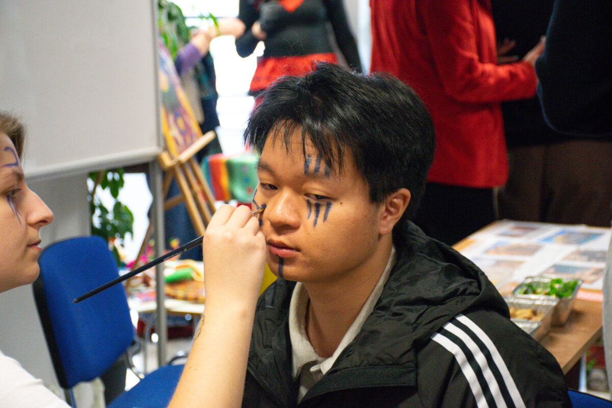 Młody mężczyzna siedzący na krześle, z czarnymi, krótkimi włosami i czarnymi pasami farby na twarzy. Na czole ma trzy pionowe czarne linie, na prawym policzku trzy podobne linie, a na środku pod dolną wargą jedną pionową kreskę. Osoba obok maluje mu twarz cienkim pędzelkiem przy lewym oku. Mężczyzna ma jasną, lekko oliwkową karnację i ubrany jest w czarną kurtkę sportową z trzema białymi paskami na rękawie; pod spodem widać jasną koszulkę z kołnierzykiem. W tle jest pomieszczenie z kilkoma osobami stojącymi i stołem z różnymi rzeczami; scena wygląda jak przygotowania do charakteryzacji lub malowania twarzy na wydarzeniu.