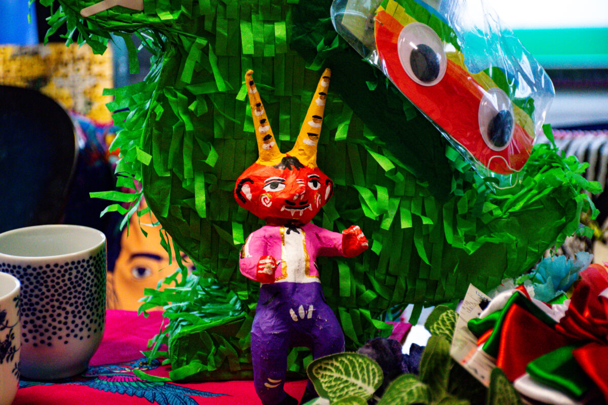 Kolorowa papierowa figurka diabełka z dwiema żółtymi rogami stoi na stole przed dużą zieloną piniatą. Ma czerwono-pomarańczową twarz z namalowanymi oczami i uśmiechem oraz mały czarny kosmyk włosów. Ubranie jest namalowane: różowa kurtka, biała koszula i fioletowe spodnie, a ręce są uniesione jak do machania lub tańca. Za figurką wypełnia kadr duża zielona piniata z pociętej bibuły. W prawym górnym rogu widać przezroczystą folię z przyczepionymi dużymi, plastikowymi „oczami” i fragment czerwonego elementu. Po lewej stronie stoją dwa kubki, jeden biały w granatowe kropki. Na dole po prawej widać zielone liście rośliny i kawałek czerwono-zielonego materiału z metką.