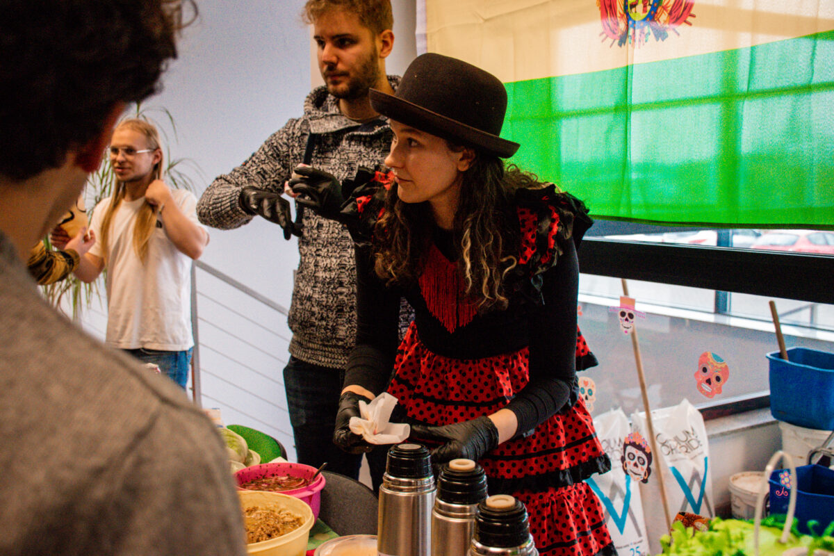 Grupa osób przy stoliku z jedzeniem w pomieszczeniu, obok dużego okna z zieloną folią lub zasłoną i dekoracjami z kolorowymi czaszkami. Na pierwszym planie jest kobieta o jasnej skórze w czarnym kapeluszu, z długimi ciemnymi włosami. Ma czarną bluzkę, czarne rękawiczki i czerwoną sukienkę w czarne kropki z falbanami; na piersi widać czerwone frędzle. Trzyma w dłoniach białą serwetkę lub chusteczkę i stoi za stolikiem. Na stole stoją trzy metalowe termosy oraz miski z jedzeniem; jedna miska wygląda na danie z ziarnami lub płatkami, obok są inne pojemniki i warzywa (w tym zielone liście). Za kobietą stoi mężczyzna o jasnej skórze w szarym, wzorzystym swetrze, w czarnych rękawiczkach, z krótkimi jasnymi włosami i zarostem. W tle po lewej jest kolejny mężczyzna o jasnej skórze z długimi blond włosami i okularami, w białej koszulce. W prawym dolnym rogu widać torby na zakupy z nadrukami oraz nieczytelny fragment napisu na jednej z nich.