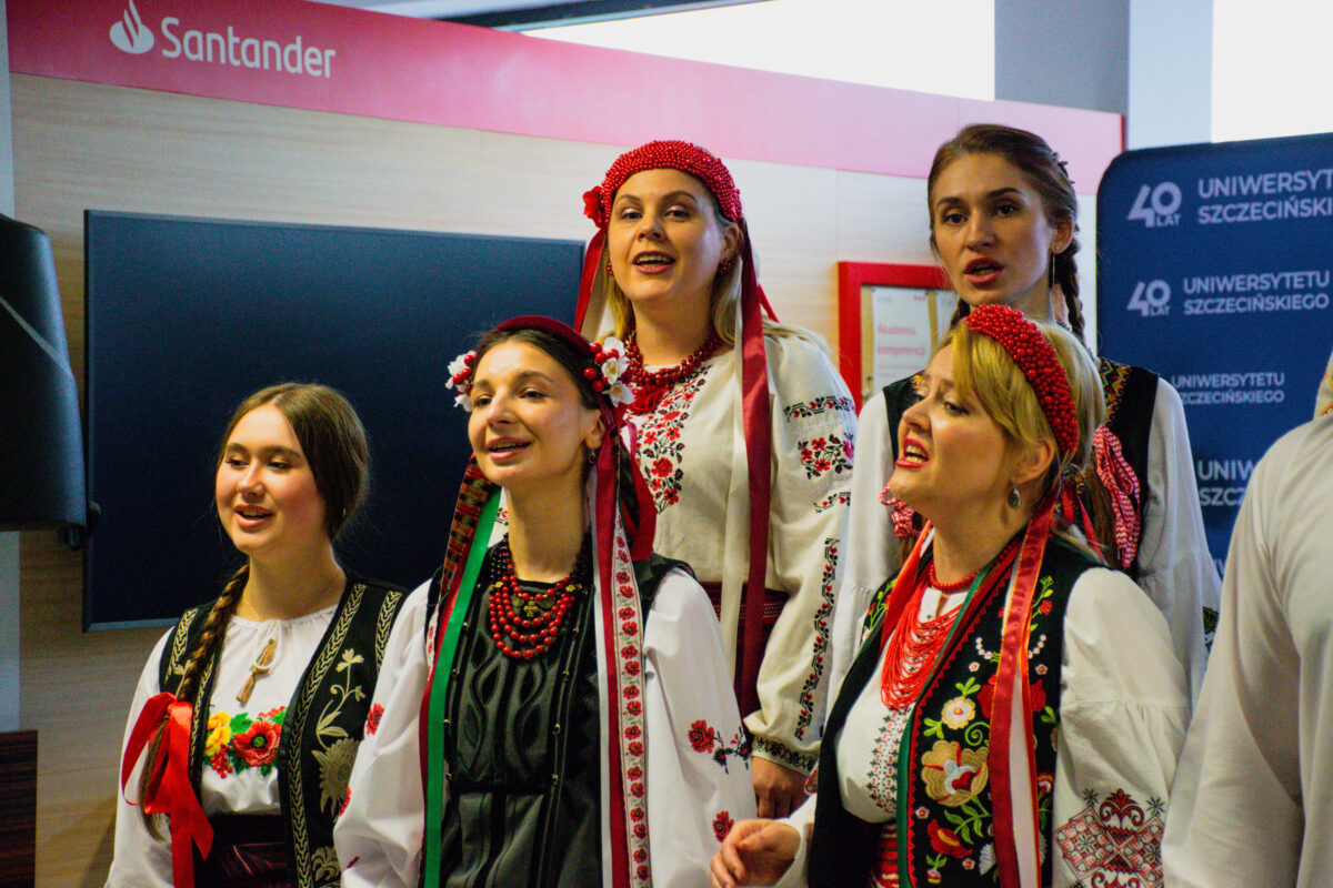 Grupa pięciu kobiet w ludowych strojach, stojących blisko siebie i śpiewających, w pomieszczeniu. Trzy kobiety na pierwszym planie mają otwarte usta, jak podczas śpiewu. Wszystkie mają jasną karnację. Ubrania to białe bluzki z haftami oraz czarne lub ciemne kamizelki/fartuchy z kolorowymi wzorami kwiatowymi. Dwie kobiety mają czerwone, koralikowe opaski lub czepce na głowie, a kilka osób ma czerwone korale i wstążki w kolorach czerwonym i zielonym. W tle widać duży czarny ekran/monitor. Na górze po lewej jest napis „Santander”. Po prawej jest granatowa ścianka z tekstem „40 LAT UNIWERSYTETU SZCZECIŃSKIEGO” (powtarzającym się).