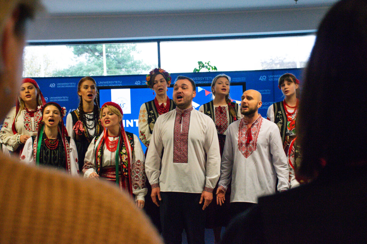 Grupa śpiewających osób w strojach ludowych na scenie, widziana z perspektywy publiczności. Na pierwszym planie stoją dwaj mężczyźni w białych koszulach z czerwonym haftem przy kołnierzu i na piersi; obaj śpiewają, usta są otwarte, ręce opuszczone wzdłuż ciała. Za nimi i obok nich stoi kilka kobiet w tradycyjnych strojach: białe bluzki, ciemne kamizelki lub gorsety, czerwone korale, kolorowe wstążki; część ma czerwone nakrycia głowy lub wianki. W tle jest niebieska ścianka z powtarzającymi się napisami „Uniwersytetu Szczecińskiego” oraz dużą cyfrą „40”. U góry widać okna z jasnym światłem dziennym. Po bokach kadru widać rozmyte sylwetki widzów.