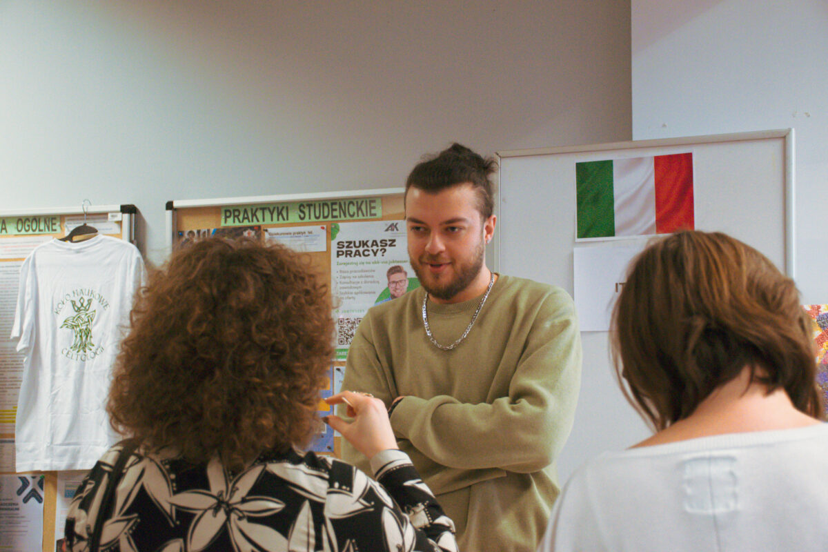 Grupa trzech osób rozmawiających w pomieszczeniu przy tablicach ogłoszeń. Mężczyzna na środku ma jasną skórę, ciemne włosy związane z tyłu i krótki zarost; ma na sobie jasnooliwkową bluzę i srebrny łańcuszek, stoi z założonymi rękami i wygląda na to, że mówi. Na pierwszym planie widać tyły dwóch osób: po lewej osoba z gęstymi, kręconymi brązowymi włosami w czarno-białym wzorzystym ubraniu, z uniesioną ręką w geście; po prawej osoba z krótkimi brązowymi włosami w jasnej bluzce. W tle są tablice ogłoszeń i plakaty. Widoczny napis „PRAKTYKI STUDENCKIE” oraz plakat z dużym tekstem „SZUKASZ PRACY?”. Po lewej wisi biała koszulka z nadrukiem. Po prawej na tablicy jest flaga Włoch.